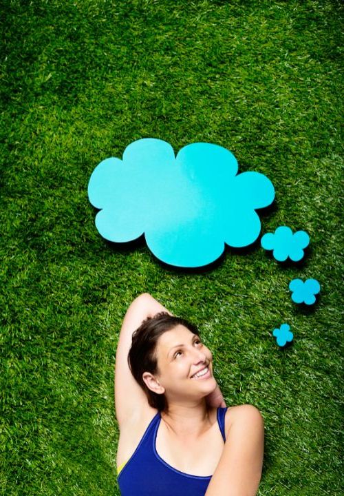 A woman practicing holistic therapy by lying on the grass with a blue thought bubble above her head. atma acupuncture