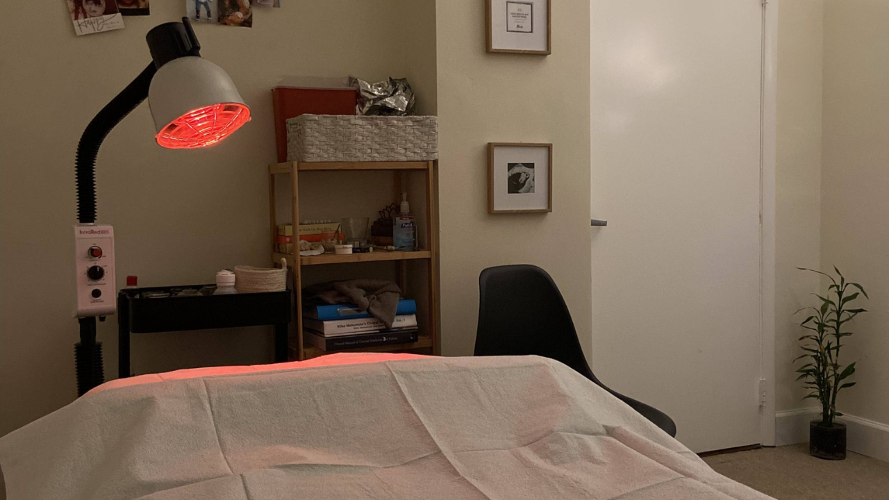 A dimly lit room with a therapy table under a red heat lamp, surrounded by shelves with various items and a small potted plant on the floor. atma acupuncture