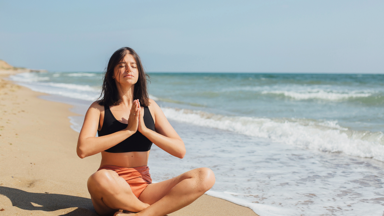 A person sits cross-legged on a sandy beach, eyes closed, hands in a prayer position, with waves in the background under a clear sky. atma acupuncture