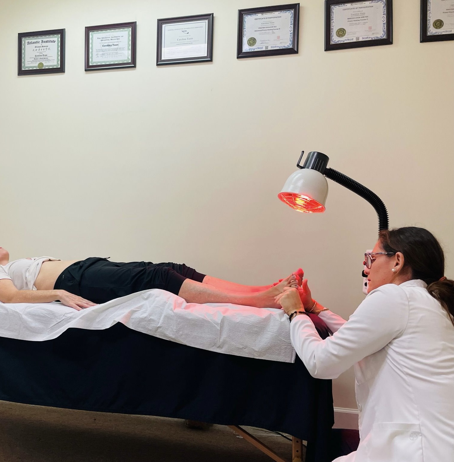 A practitioner in a white coat massages a patient's feet under a red heat lamp in a treatment room with certificates on the wall. atma acupuncture