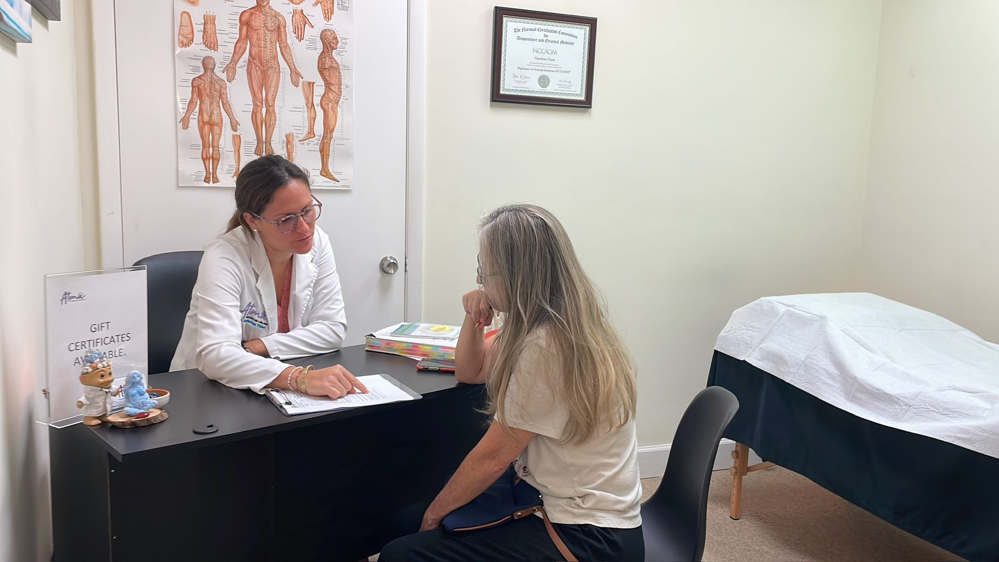 A healthcare professional in a white coat consults with a seated patient in a medical office with anatomical charts, certificates, and an exam table visible. atma acupuncture